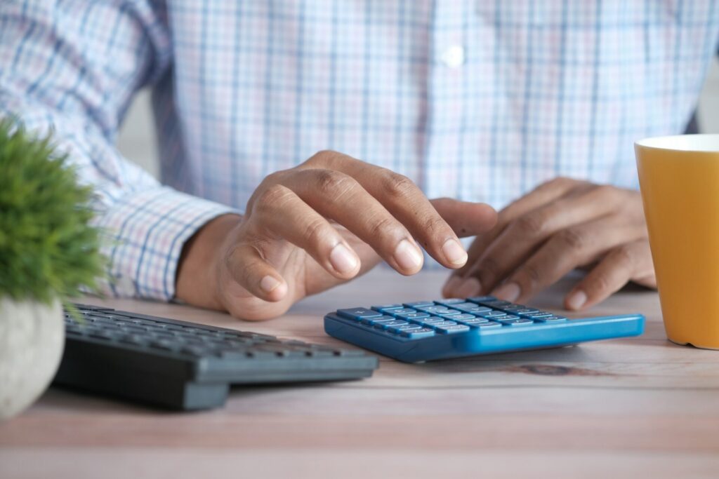 A person in a checked shirt taps a blue calculator beside a keyboard and a yellow mug