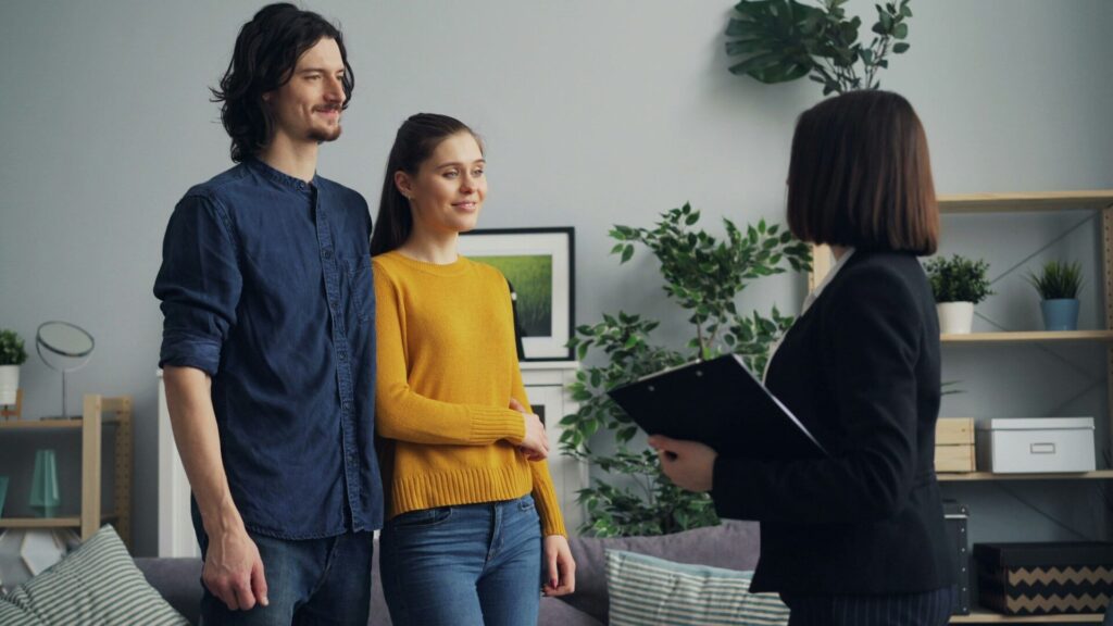 A real estate agent with a clipboard talks with a young couple in a living room