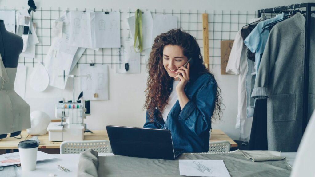 A clothing designer talks on the phone at a laptop in her studio