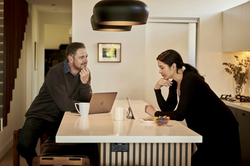 A man at a laptop and a woman with a tablet sit across a kitchen island, each resting a chin on one hand