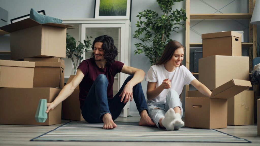 A smiling couple sits on the floor of their new home and unpacks moving boxes