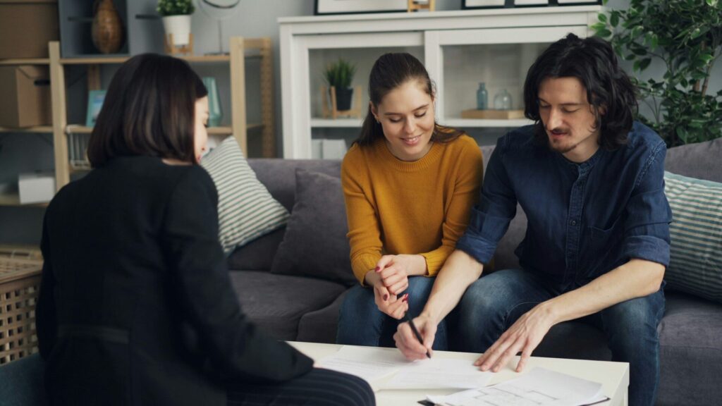 An advisor reviews paperwork with a young couple who sit on a living room sofa
