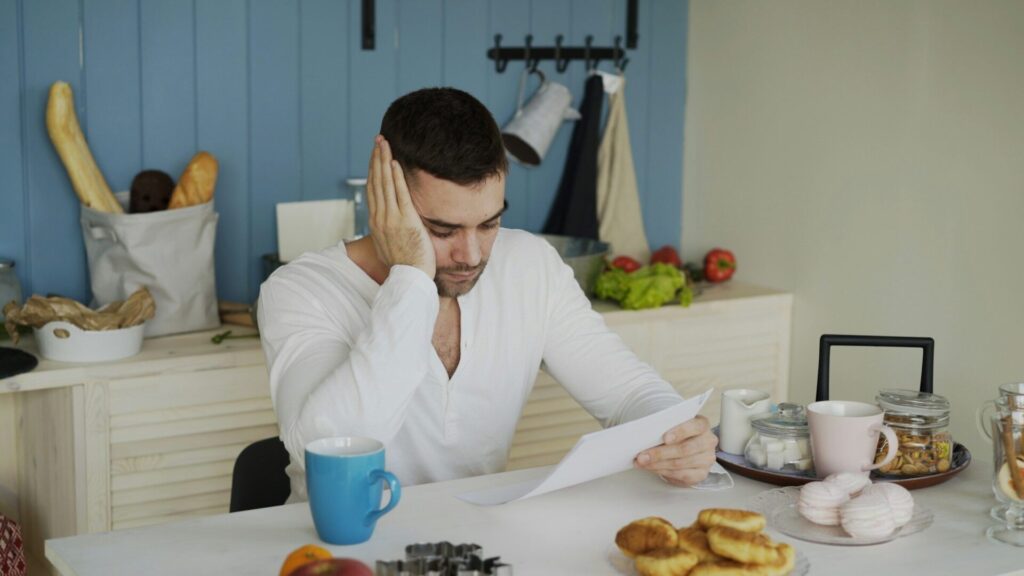 A man rests his head on his hand while reading a letter at his kitchen table