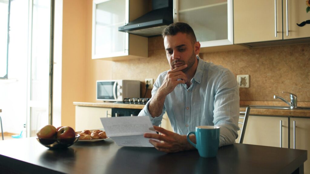 A man in a light blue shirt reads a letter at his kitchen table, chin in hand, next to a blue mug and a bowl of fruit