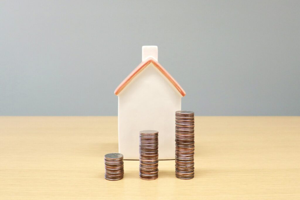 A small white model house with a coral roof stands behind three stacks of coins that grow taller from left to right