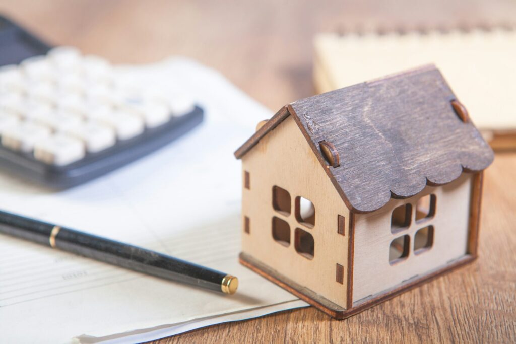 A small wooden model house sits on loan paperwork next to a pen and a calculator on a wooden desk