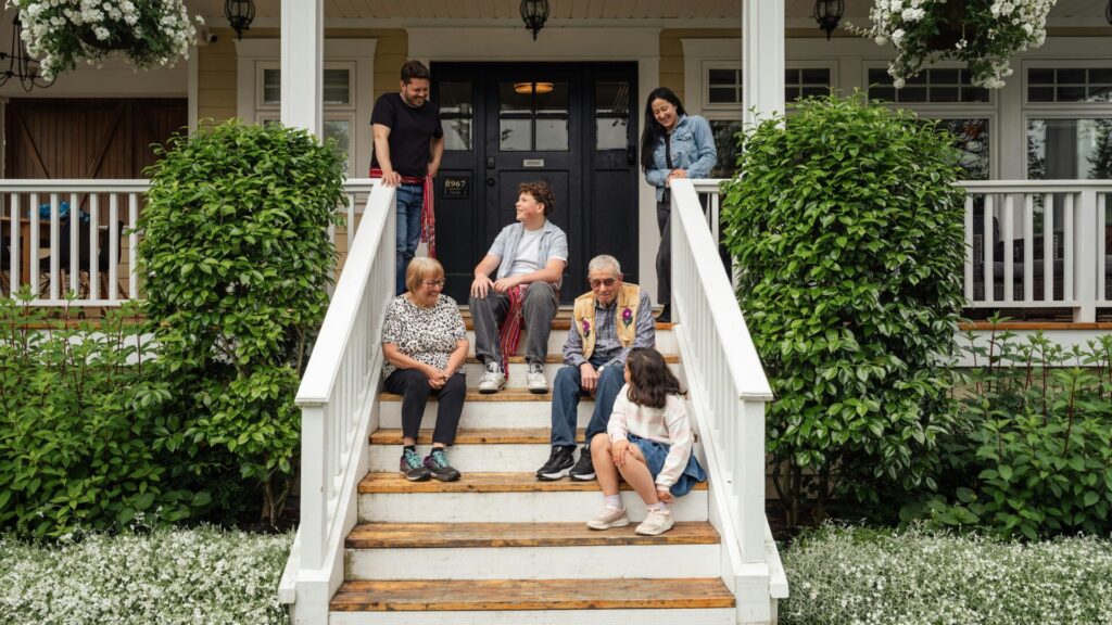 Three generations of a family relax on the front steps of a white-porched house