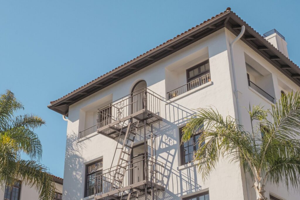 A white stucco apartment building with a tile roof, a fire escape and palm trees