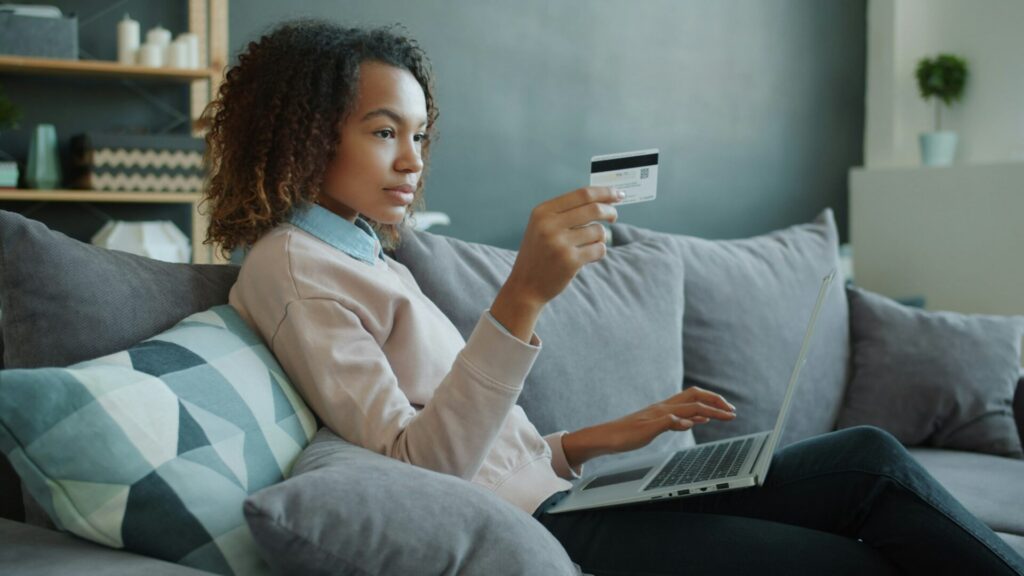A woman on a sofa holds a credit card while using a laptop